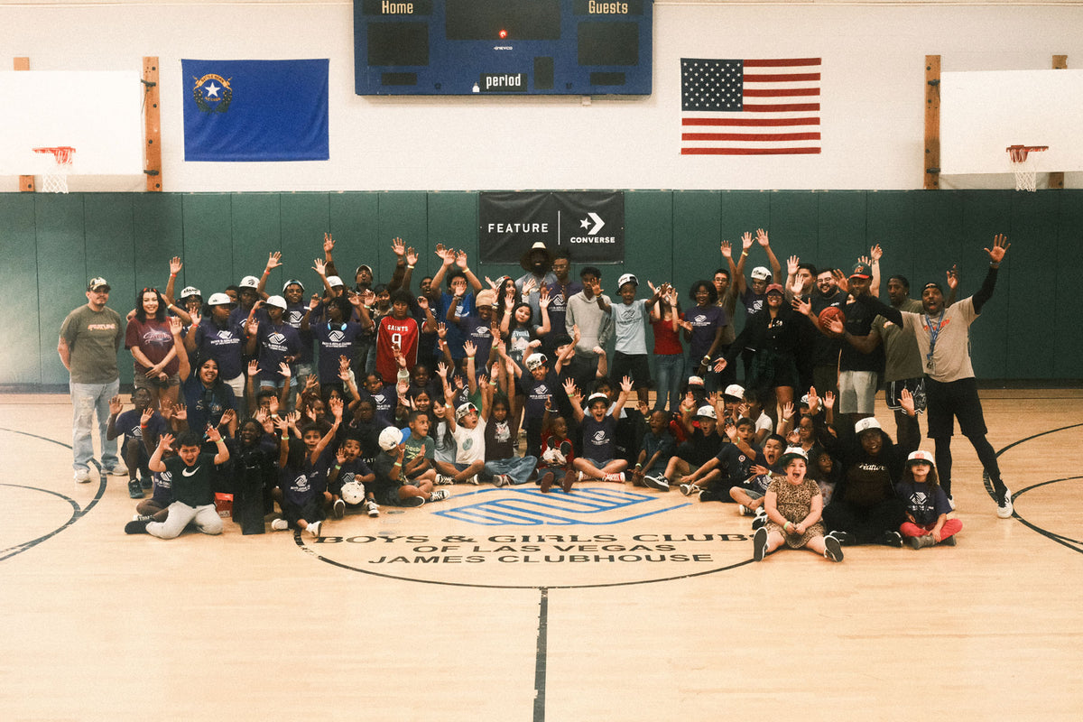 A large group of children and adults pose together on a gymnasium floor, smiling and raising their hands. The group is gathered for an event at the Boys & Girls Club of Las Vegas, with banners for FEATURE and Converse hanging in the background.